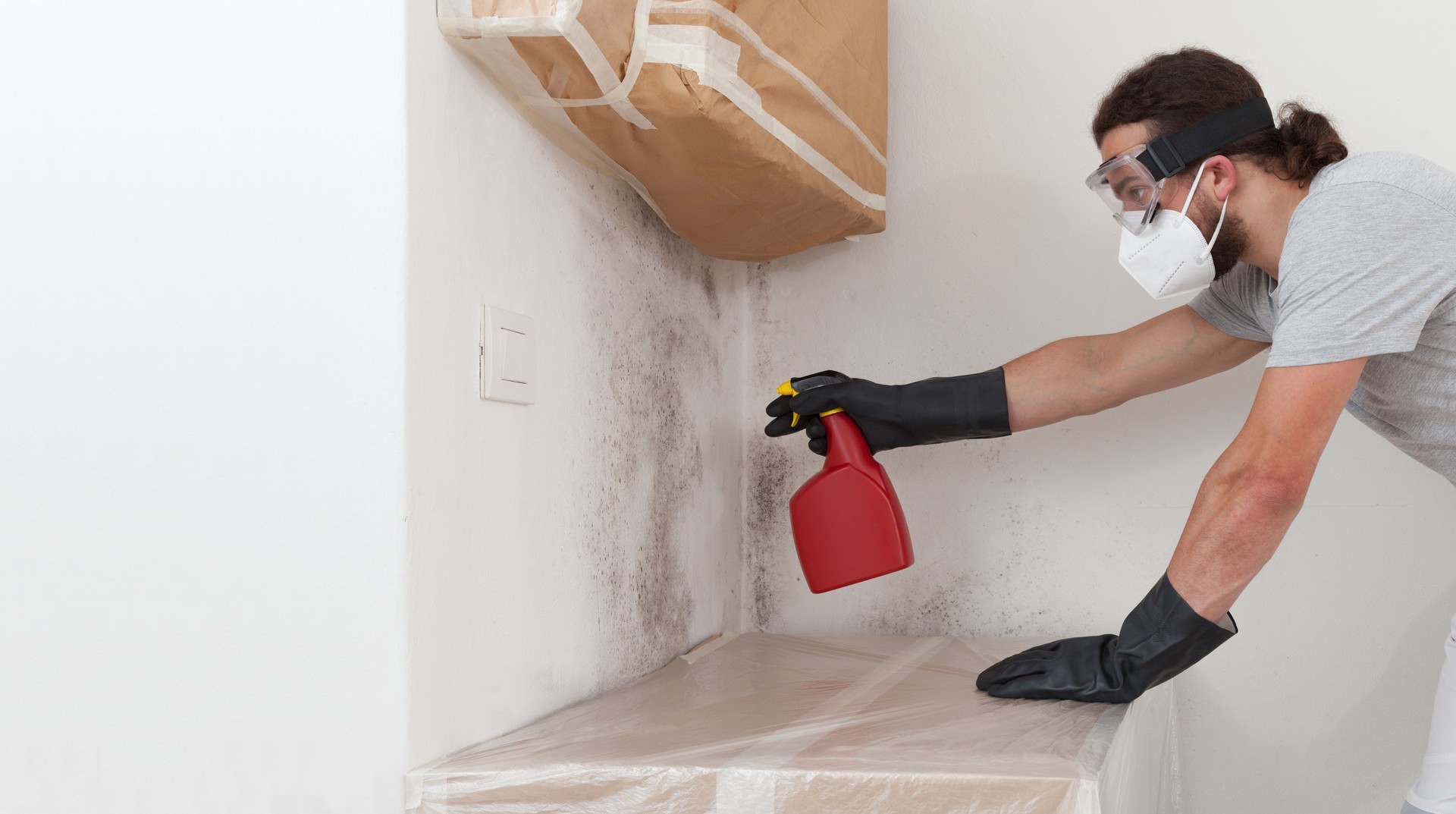 Man worker spraying product on wall to remove mold, wearing protective gloves and a respirator mask, using a spray bottle. Remediation of moldy wall before house painting at home renovation site